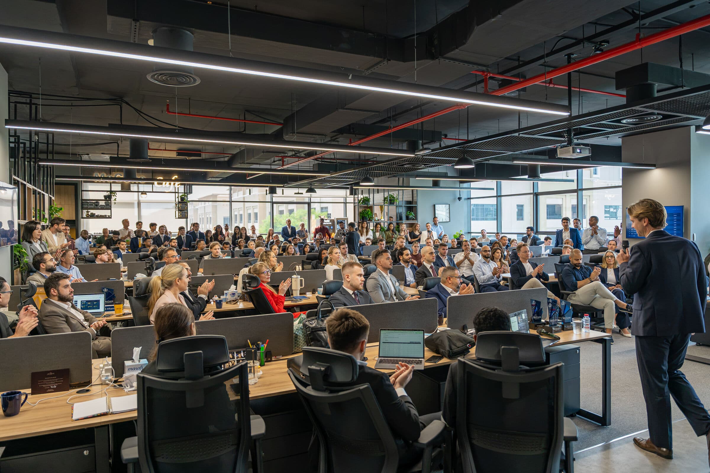 Sam McCone, microphone in hand, addresses a packed open-plan office of more than a hundred McCone Properties agents, who sit at their desks and stand along the back wall, applauding.